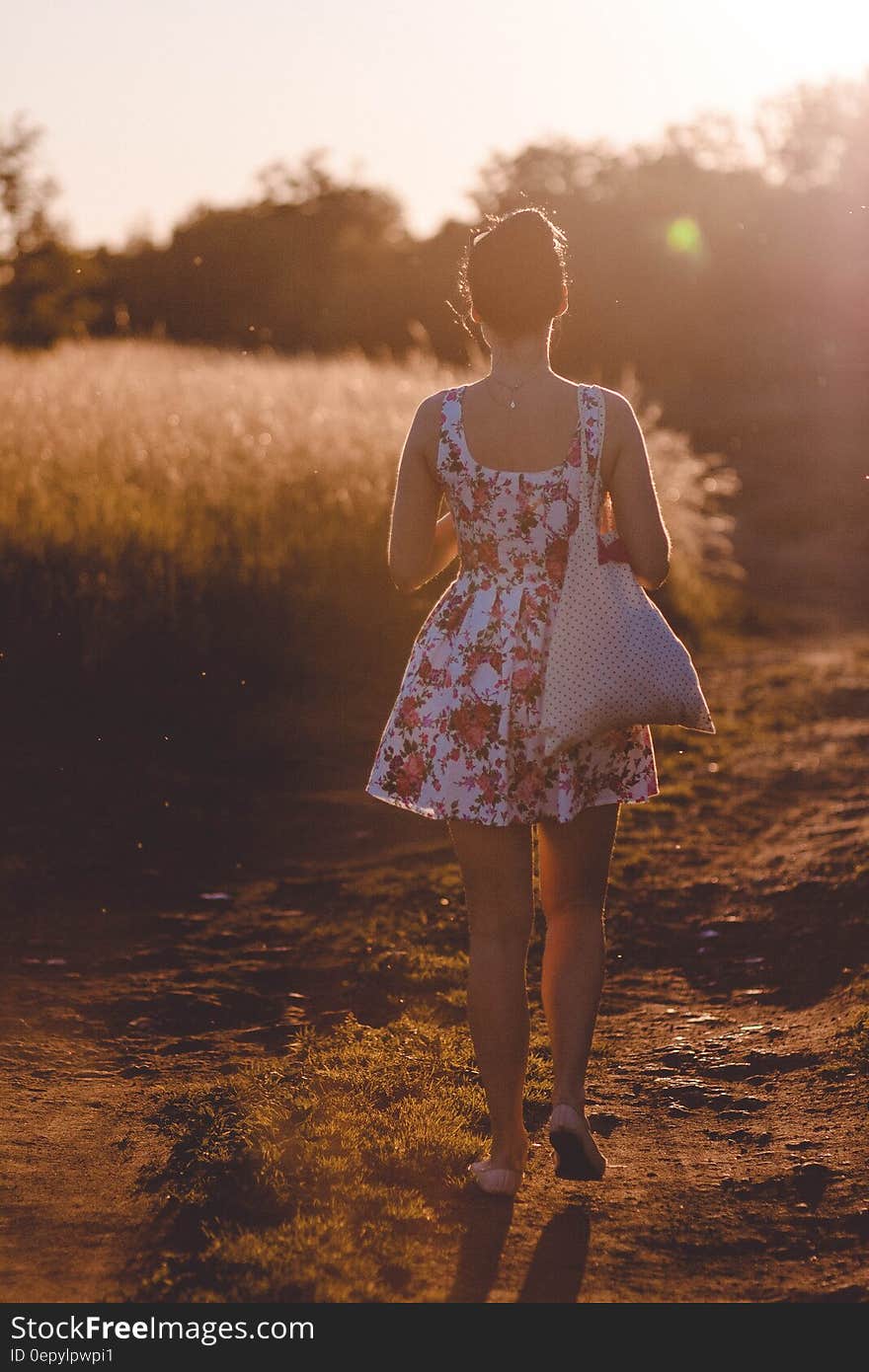 Woman in White and Pink Floral Sleeveless Dress Walking on Brown Road during Sunlight