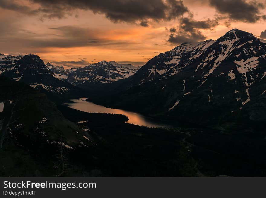 Sunset over mountain lake with snowy peaks and cloudy skies.