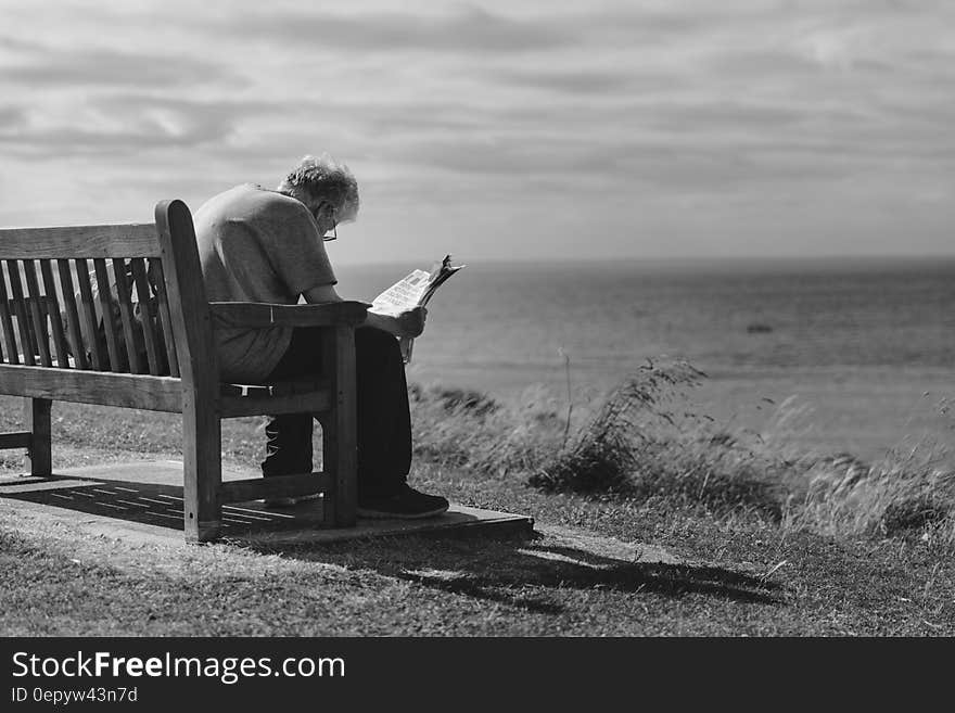 Grayscale Photo of Man Sitting on Brown Wooden Bench Reading News Paper during Day Time