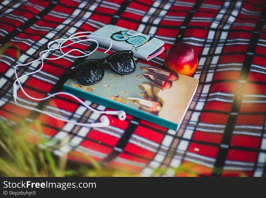 A plaid blanket with a book, smartphone with headphones, sunglasses and a fruit. A plaid blanket with a book, smartphone with headphones, sunglasses and a fruit.