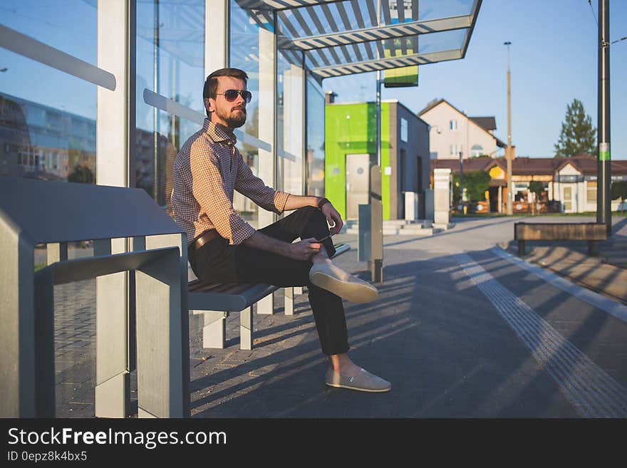 Man Wearing Sunglasses Sitting at Bus Stop during Daytime