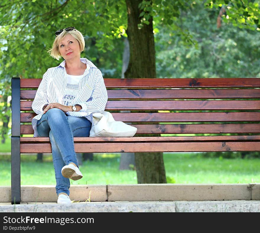 Thinking Woman in White Jacket and White Scoop Neck Shirt Blue Denim Jeans Sitting on Brown Wooden Bench Beside Green Trees during