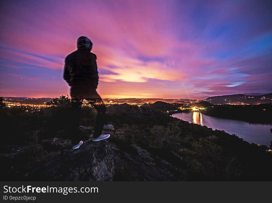 Man standing on rocky cliff wearing hoodie jacket over river at sunset. Man standing on rocky cliff wearing hoodie jacket over river at sunset.