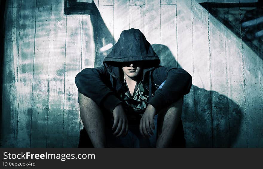 Person Wearing Black Zip Hoodie Sitting in Front of Gray Wooden Plank Wall during Nighttime