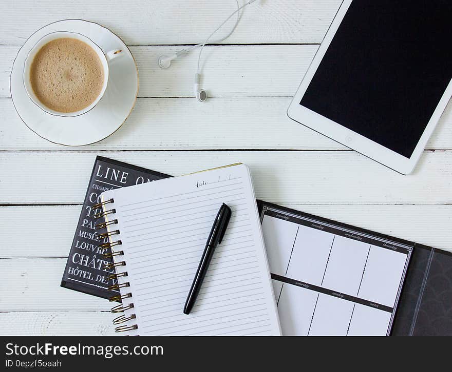 Black Pen on White Writing Spring Notebook Between White Ipad and White Ceramic Mug With Latte on White Plate