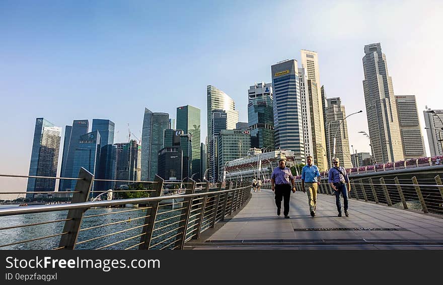 Three Men Walking on Bridge in Front of High Rise Buildings at Daytime