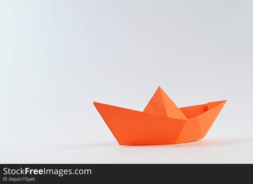 An orange colored stylized boat made by origami, that is folding plain orange paper, white background. An orange colored stylized boat made by origami, that is folding plain orange paper, white background.