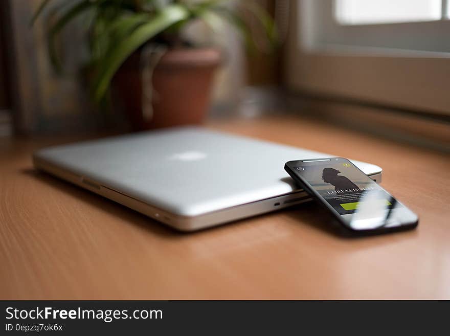 Smartphone Beside Silver Macbook on Brown Wooden Table With Potted Plant in the Background