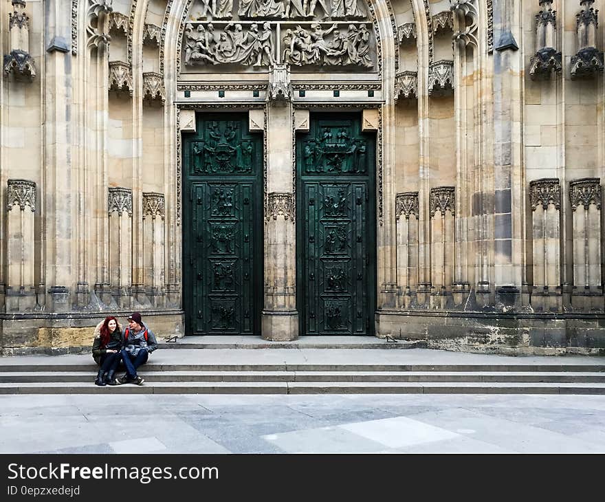 Couple Sitting on Ladders Outside Yellow Concrete Building With 2 Green Wooden Doors