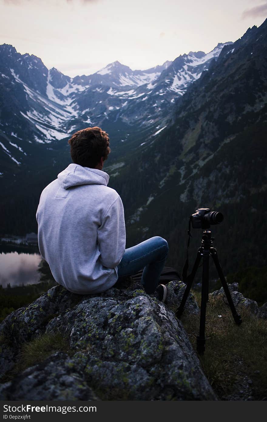 Man in Gray Hoodie Sitting on Gray Stone With Dslr Camera Tripod