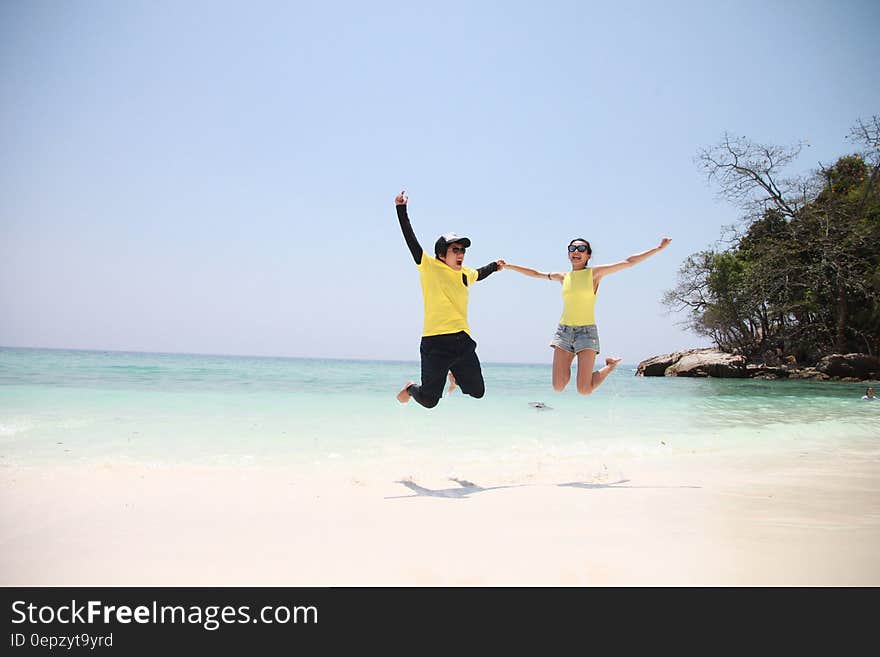 Woman in Blue Denim Mini Short Smiling While Holding Another Person in a Jump Shot Photo at Seashore during Daytime