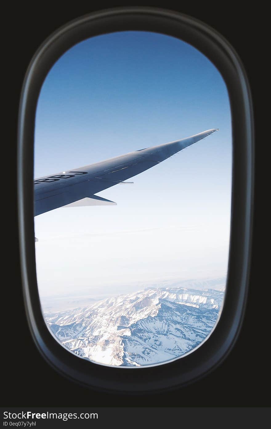 A view of an airplane wing from the airplane window and mountains covered in snow down below.