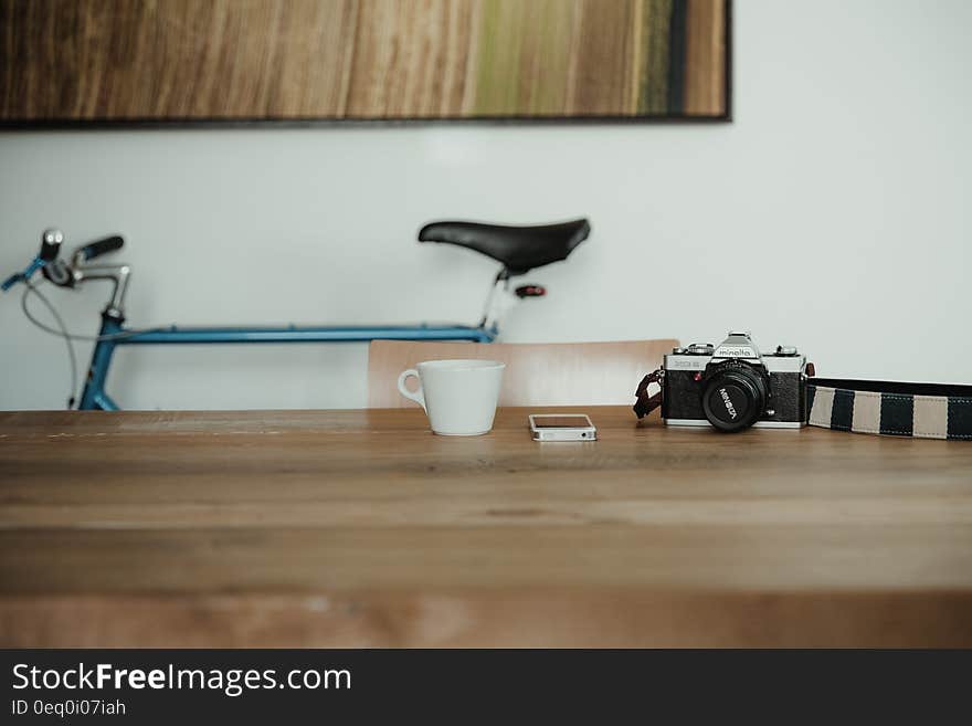 Camera and smartphone with white china mug on wooden desk inside room with bicycle. Camera and smartphone with white china mug on wooden desk inside room with bicycle.