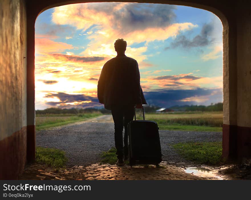 Man Pulling Luggage Walking Near Gray Concrete Road during Sunset