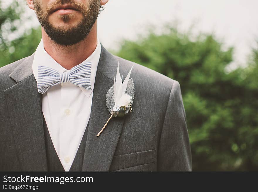 Closeup of the groom with beard and mustache showing white shirt, bow tie, white flower (lily), light gray suit, background of green trees and pale sky.