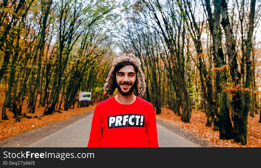 Man wearing red sweatshirt and fur hat standing in country road lined with autumn trees on sunny day. Man wearing red sweatshirt and fur hat standing in country road lined with autumn trees on sunny day.