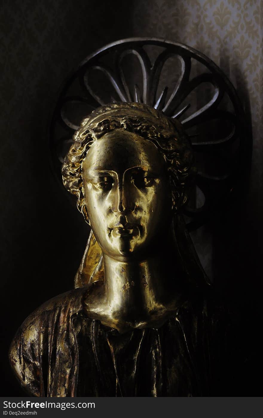 Sculpture of an angel with hair band and robes finished in gold with a dark halo, and mainly dark background.