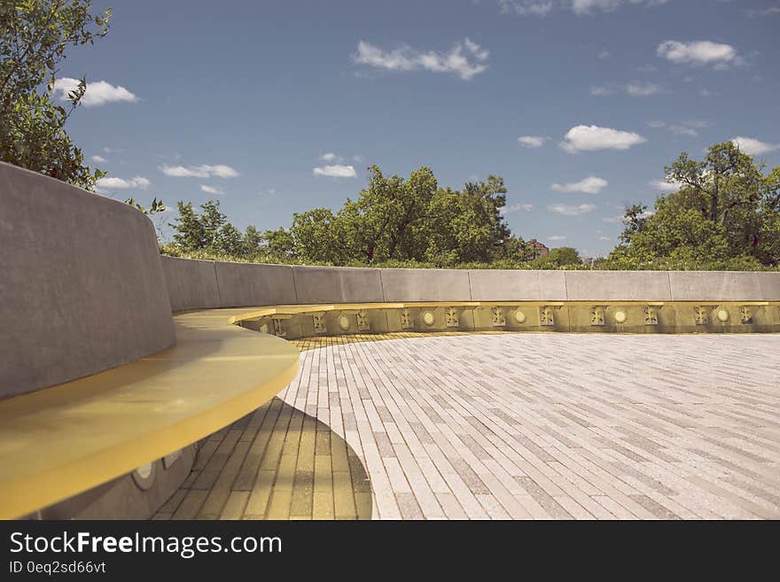 Long curved bench in city park with trees, blue sky and clouds in background.