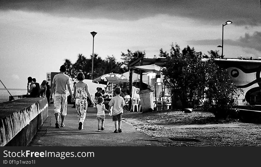 Family, mother, father and two children holding hands and walking at the seaside with boys fishing off the sea wall and a cafe and fairground style entertainment on the right. Family, mother, father and two children holding hands and walking at the seaside with boys fishing off the sea wall and a cafe and fairground style entertainment on the right.