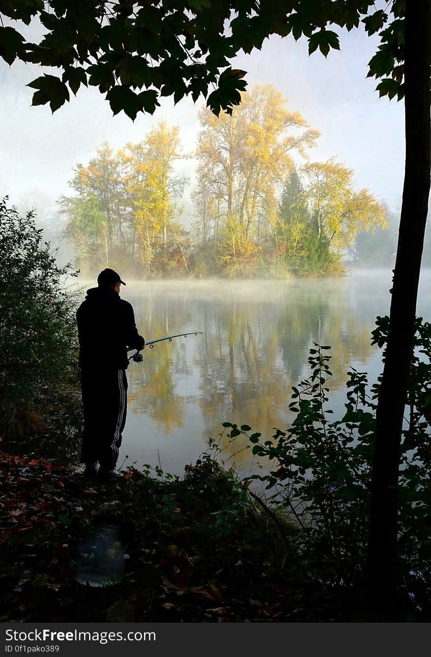 Silhouette of fisherman fishing in tranquil lake or river framed by tree and bushes. A magical sunlit island in the river gives 3D impact. Silhouette of fisherman fishing in tranquil lake or river framed by tree and bushes. A magical sunlit island in the river gives 3D impact.