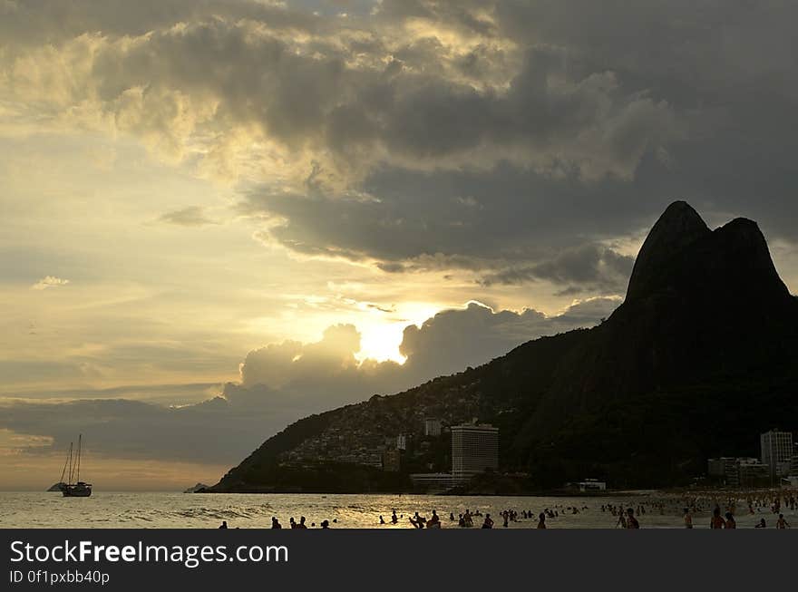 Sailboats off rocky cliff along coastline at sunset in cloudy skies.