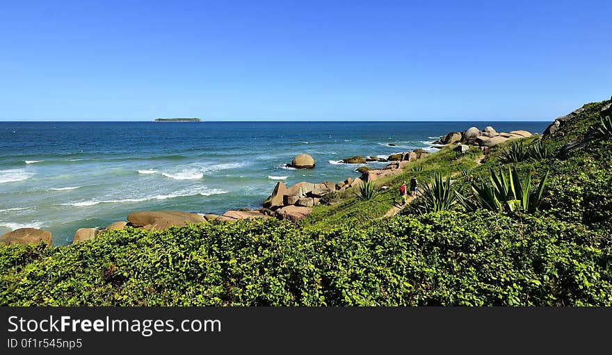 Green hillside down to rocks along blue waters of shore against blue skies on sunny day. Green hillside down to rocks along blue waters of shore against blue skies on sunny day.