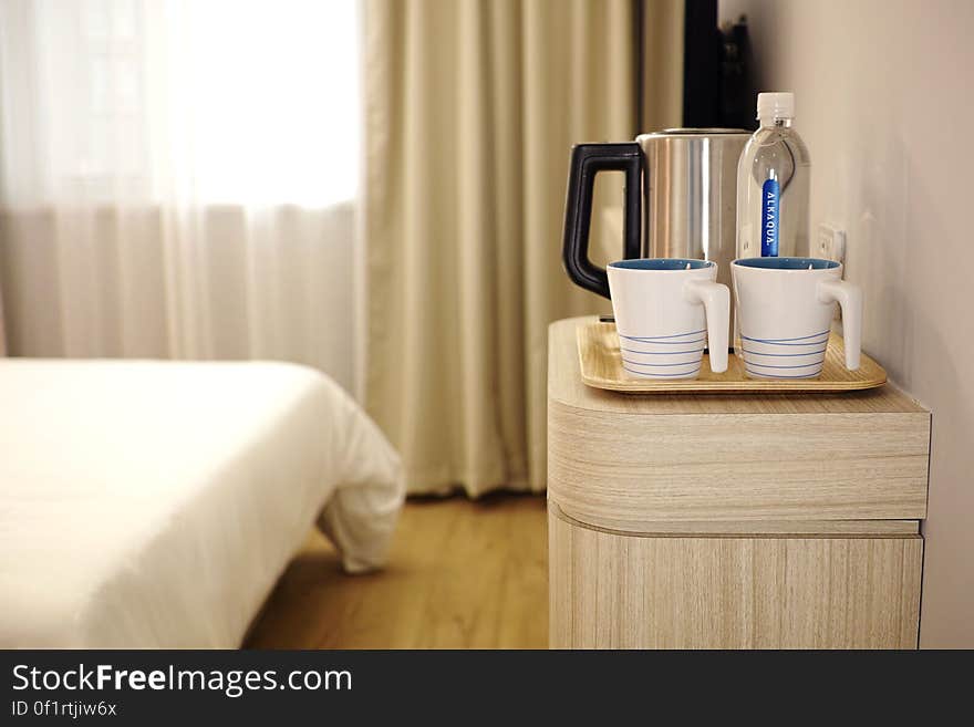 A hotel or guest room with a water kettle, cups and other items on the bedside table. A hotel or guest room with a water kettle, cups and other items on the bedside table.