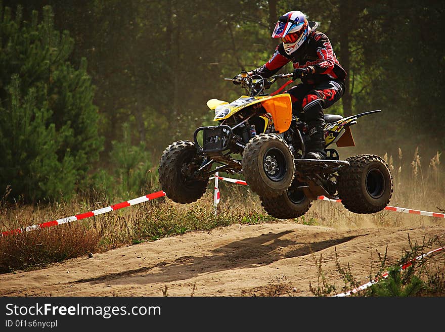 Quad bike with rider in full helmet and black and red suit taking off at hump in the race track throwing up dust, background of green forest. Quad bike with rider in full helmet and black and red suit taking off at hump in the race track throwing up dust, background of green forest.