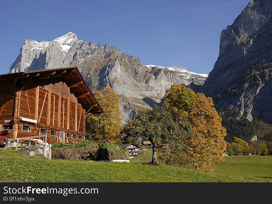 Wooden chalet with exposed beams, and manicured garden with trees and towering over the roof a range of high mountains and blue sky. Wooden chalet with exposed beams, and manicured garden with trees and towering over the roof a range of high mountains and blue sky.