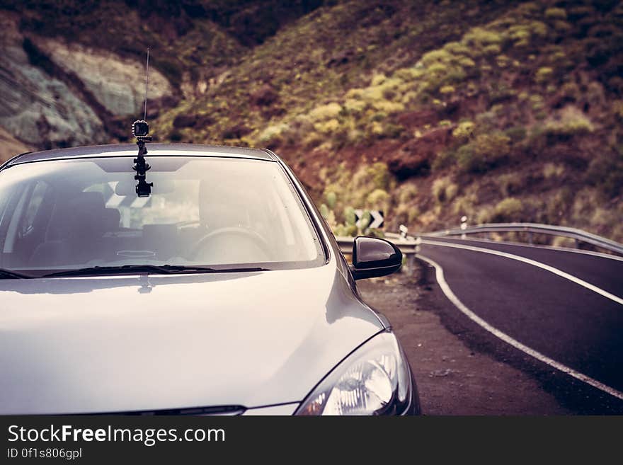 Silver saloon car with small camera fixed to the roof parked beside a narrow road with white markings through wild mountainous country. Silver saloon car with small camera fixed to the roof parked beside a narrow road with white markings through wild mountainous country.