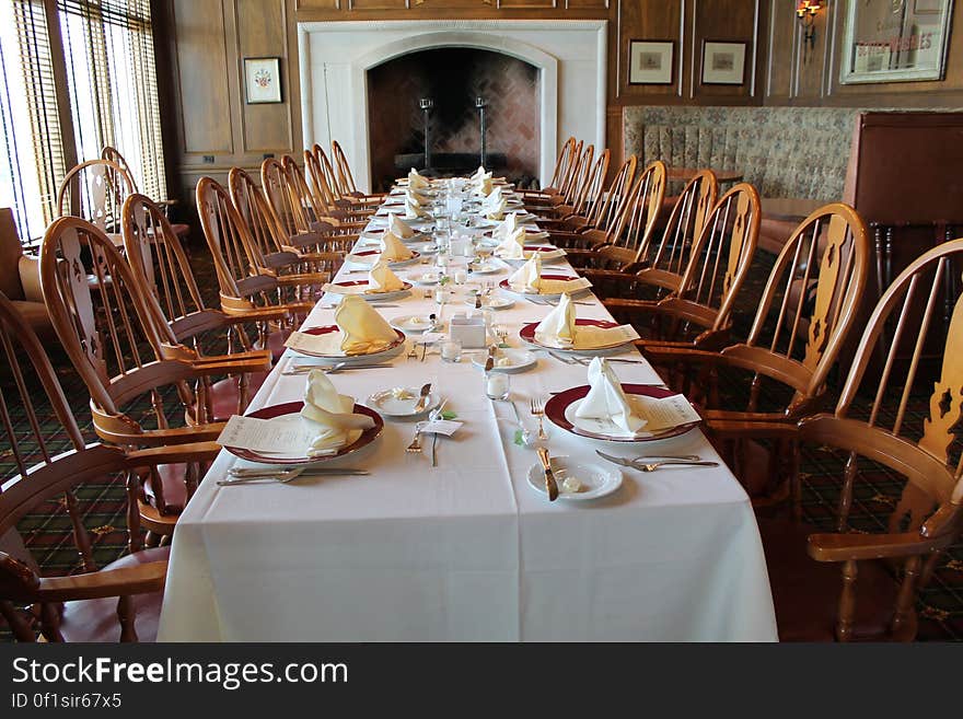 A long dining room table with a white tablecloth and formal place settings, ten chairs on each side, ending in front of a large fireplace. A long dining room table with a white tablecloth and formal place settings, ten chairs on each side, ending in front of a large fireplace.