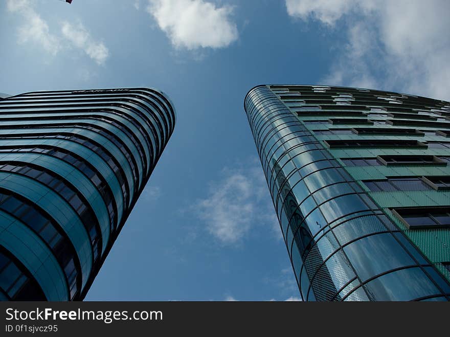 View upwards beside and between two adjacent skyscrapers built mainly of glass and metal, (looking almost identical), background of blue sky and white cloud.