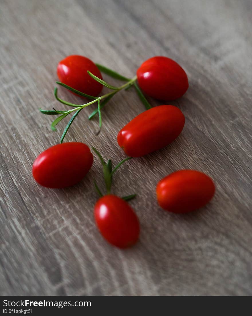 Six small cherry or plum tomatoes together with a sprig of rosemary, lying on a grainy wooden table top. Six small cherry or plum tomatoes together with a sprig of rosemary, lying on a grainy wooden table top.