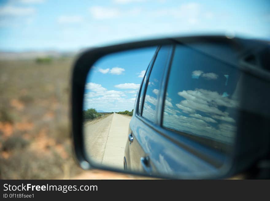 View through car's side mirror showing road, sky, cloud and car windows with blurred view of surrounding countryside. View through car's side mirror showing road, sky, cloud and car windows with blurred view of surrounding countryside.