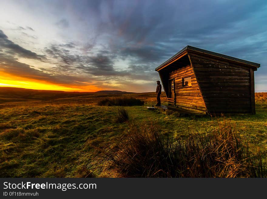 Solitary figure standing beside a log cabin on the moors at sunset with a yellow and orange sky.