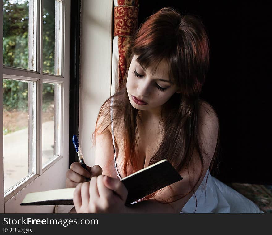 Portrait of pretty young girl with long red hair writing in her diary while seated next to a window. Portrait of pretty young girl with long red hair writing in her diary while seated next to a window.