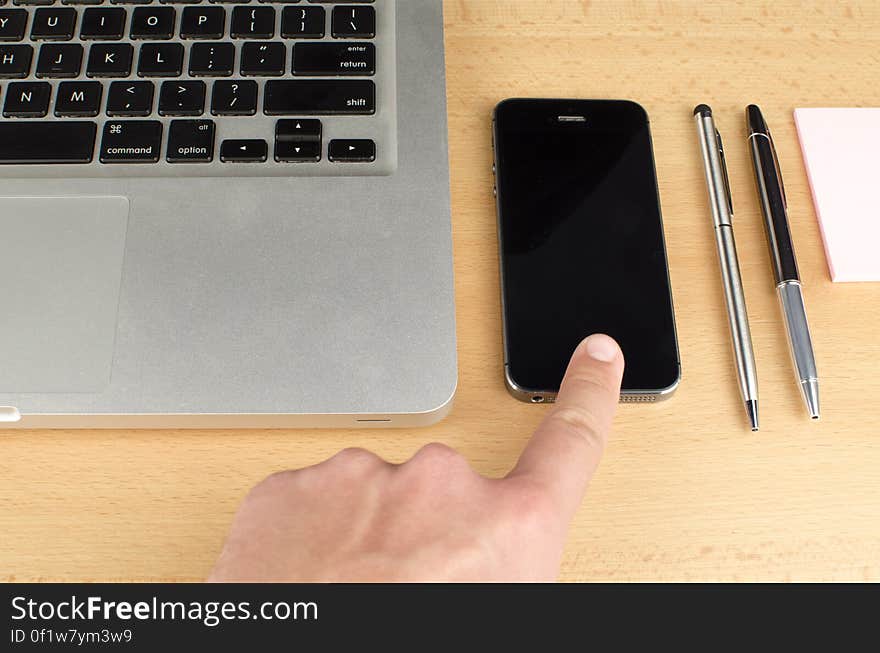 A finger touching a smartphone, laptop keyboard and pens next to it. A finger touching a smartphone, laptop keyboard and pens next to it.