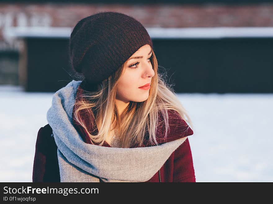 Portrait of young blond woman in dark brown beanie hat (tea cozy hat) and red jacket with cold snowy background. Portrait of young blond woman in dark brown beanie hat (tea cozy hat) and red jacket with cold snowy background.