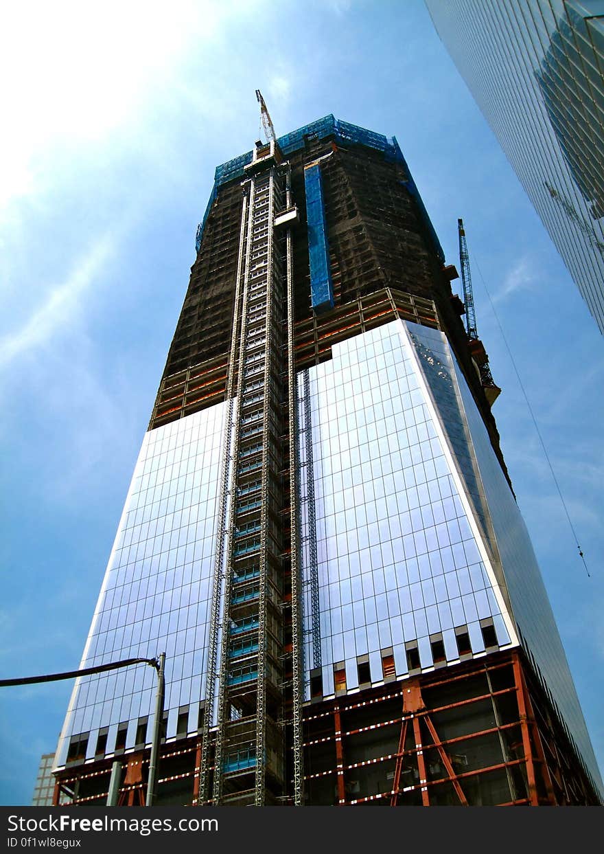 Skyscraper building under construction with ladder (or lift) on the outer wall and heavy lift crane on the far side, blue sky and thin cloud background. Skyscraper building under construction with ladder (or lift) on the outer wall and heavy lift crane on the far side, blue sky and thin cloud background.