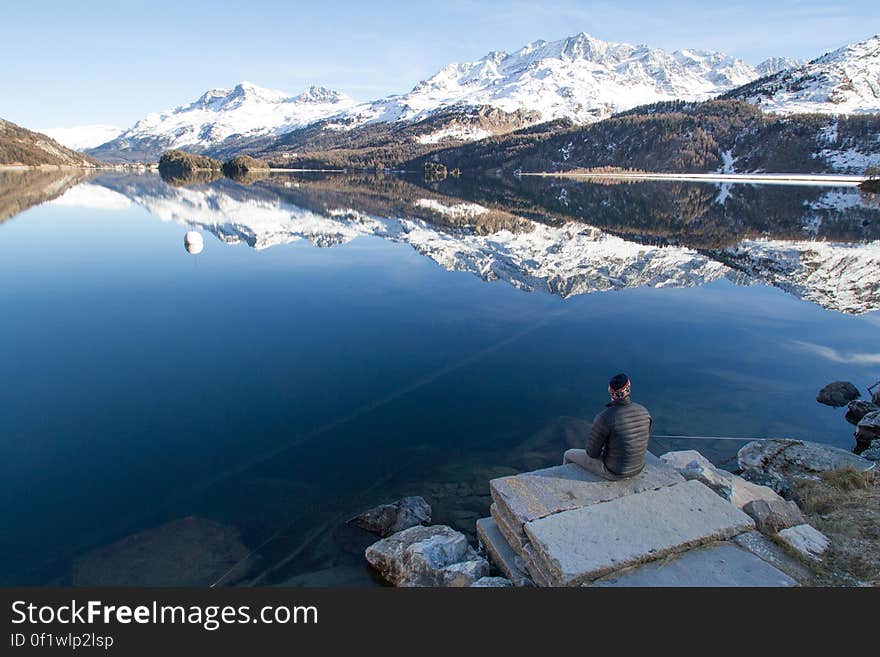 A man sitting by a smooth, calm lake with snow covered mountains reflecting in the water. A man sitting by a smooth, calm lake with snow covered mountains reflecting in the water.