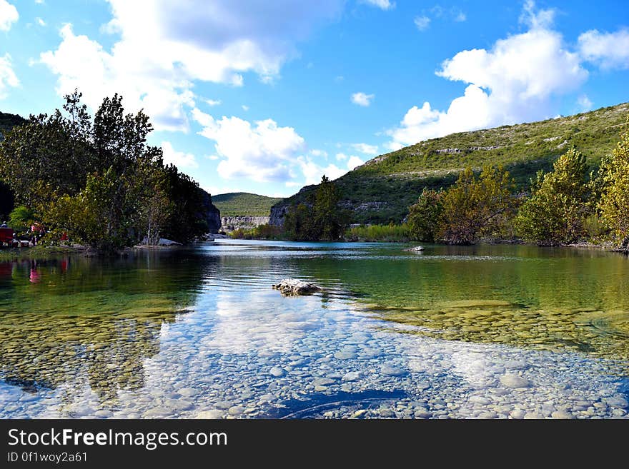 Shallow river flowing over large pebbles in the river bed with hills on both sides covered mostly with trees, blue sky and fluffy clouds. Shallow river flowing over large pebbles in the river bed with hills on both sides covered mostly with trees, blue sky and fluffy clouds.