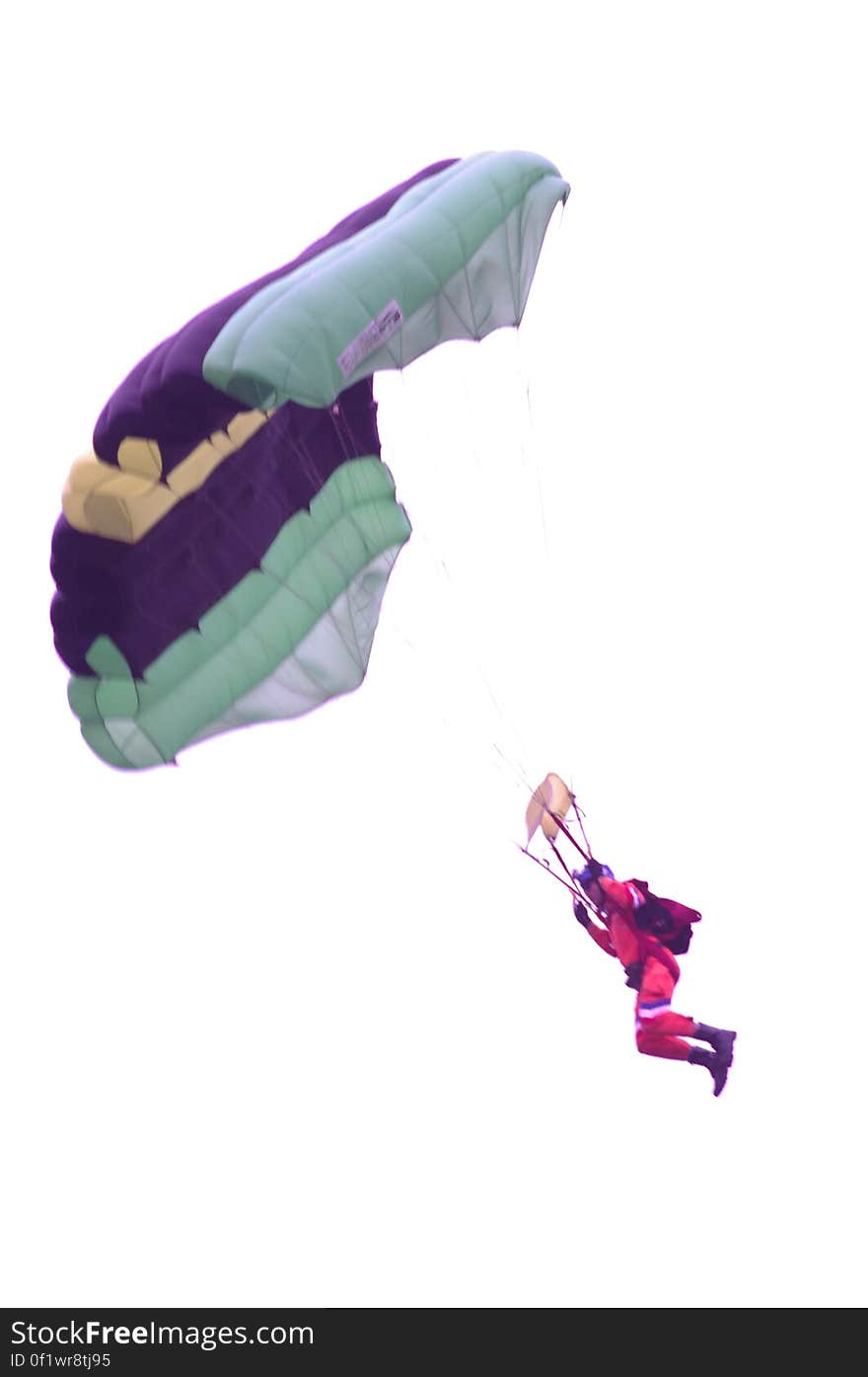 Man in purple suit and black boots with reserve parachute on his back descending while attached to a large voluminous purple, gray , green and beige colored parachute, white sky background. Man in purple suit and black boots with reserve parachute on his back descending while attached to a large voluminous purple, gray , green and beige colored parachute, white sky background.