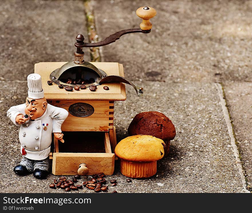 A hand crank coffee bean grinder with muffins and a baker figurine. A hand crank coffee bean grinder with muffins and a baker figurine.