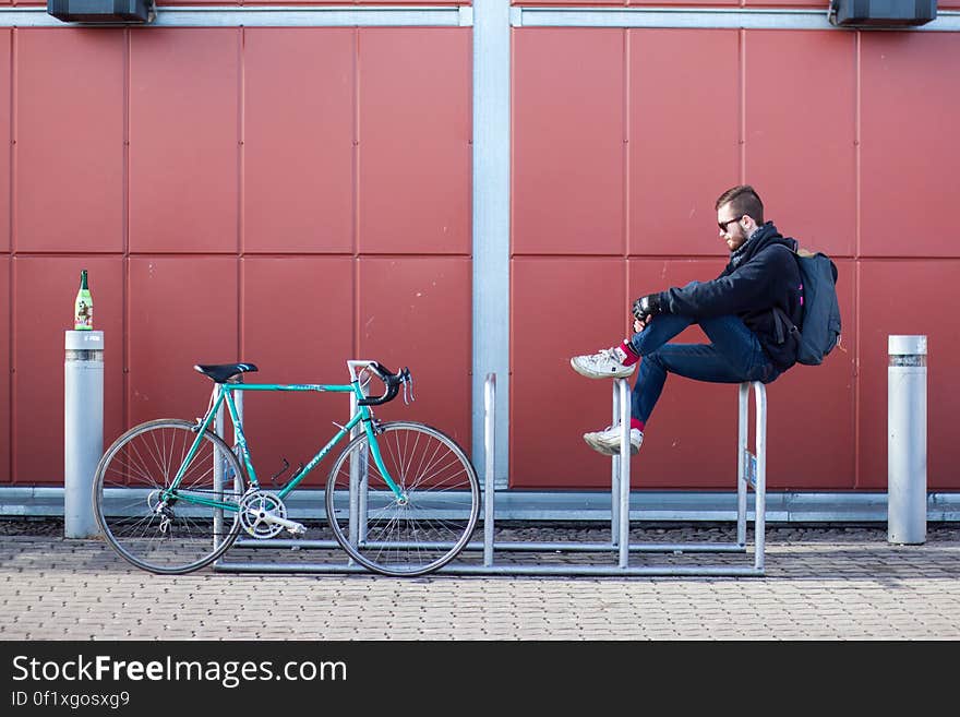 A man sitting on a bike parking rack with a bike parked next to him. A man sitting on a bike parking rack with a bike parked next to him.