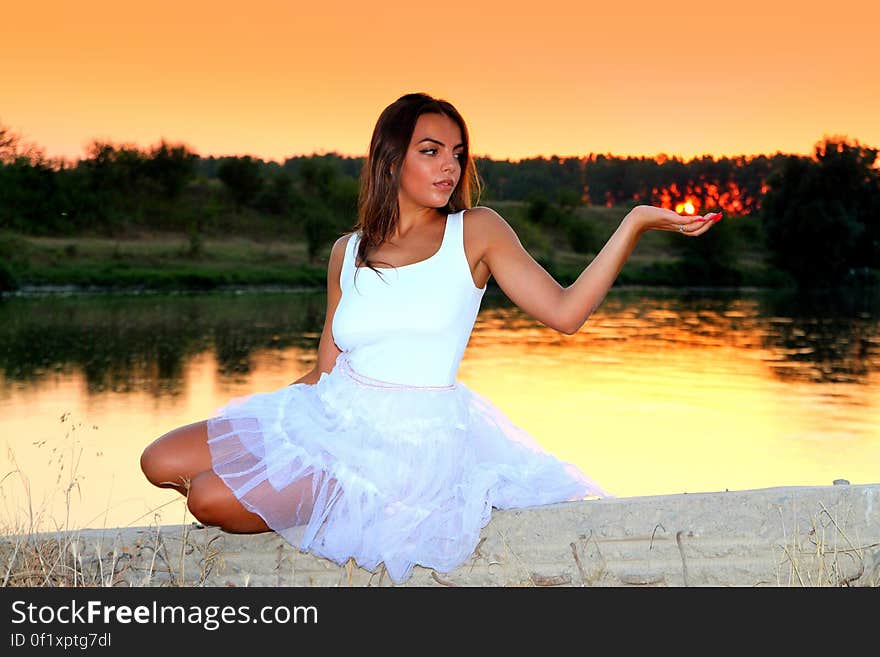 Woman in White Square Neckline Sleeveless Dress Sitting on Beige Wall Beside Body of Water during Golden Time