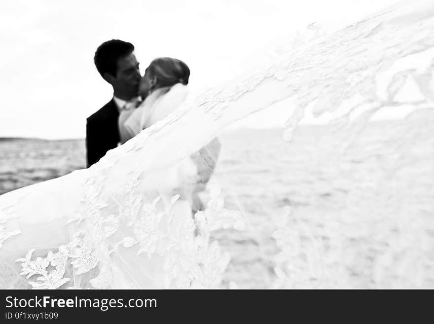 Bride and groom kissing on the sea shore while she is still wearing her dazzling white wedding dress and he a dark formal suit.