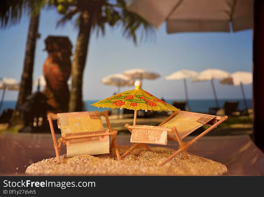 A close up of a miniature model of a parasole and two sunbeds on sand, tropical beach in the background. A close up of a miniature model of a parasole and two sunbeds on sand, tropical beach in the background.