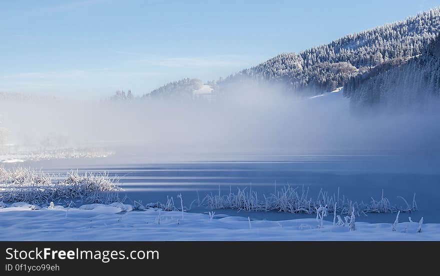 A frozen lake with fog and hills with forests.