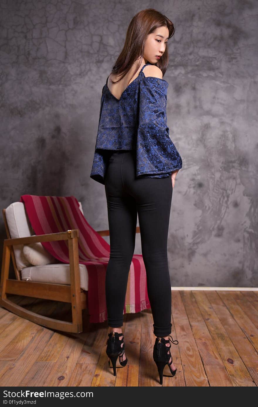 Attractive young Asian girl (Japanese) posing while wearing blue top, black tight fitting trousers and extremely high heels. Bare room except for one easy chair and gray wall. Attractive young Asian girl (Japanese) posing while wearing blue top, black tight fitting trousers and extremely high heels. Bare room except for one easy chair and gray wall.