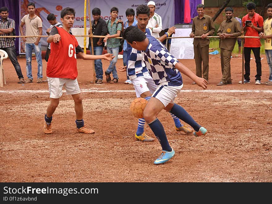 Players on dirt field with spectators during soccer match.